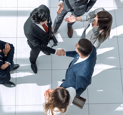 A group of business professionals in formal attire are engaged in a meeting on a tiled floor, with two men shaking hands, signaling agreement or partnership.