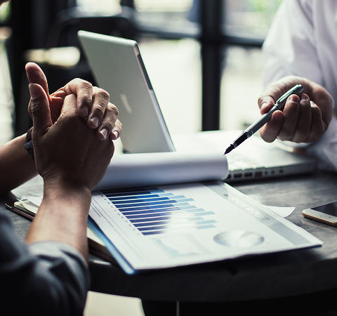 Two people are discussing over a table with business charts, a laptop, and a smartphone. The setting suggests a professional and analytical atmosphere.