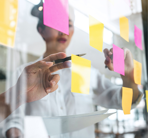 Person in a white shirt writing on colorful sticky notes on a glass wall. The scene conveys a brainstorming or planning session.