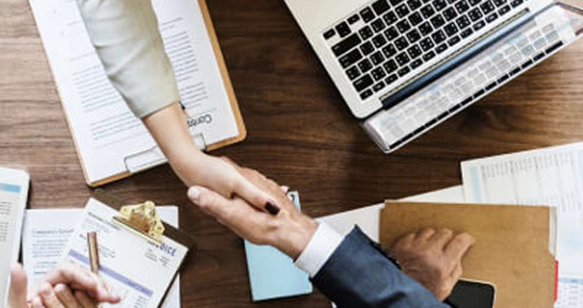 Overhead view of people shaking hands over a wooden table, surrounded by a laptop, documents, and a clipboard, conveying a business agreement.