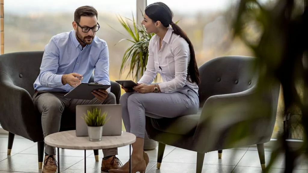 A man and woman in business attire sit in modern armchairs, discussing documents, with a potted plant nearby, in a bright office setting.