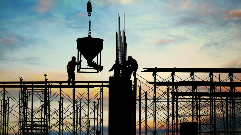 Silhouetted construction workers operate a crane at a building site against a colorful sunset sky. The scene conveys a tone of industriousness and teamwork.